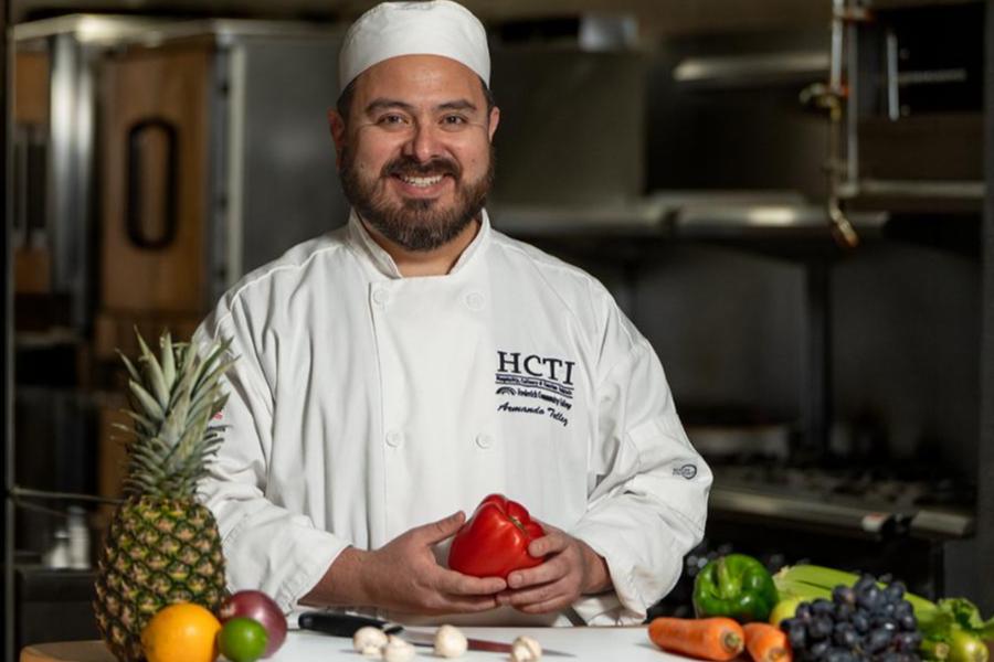 A smiling chef in an HCTI chef's coat and hat holds fresh produce in a professional kitchen.